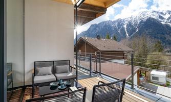 A cozy balcony with modern furniture and a view of the mountains. The sun is shining and the sky is clear.