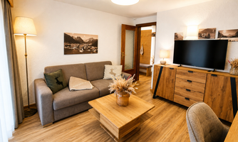 A cozy living room with a gray sofa and a wooden table. The room is stylishly decorated and has a large television as well as decorative pictures on the wall.