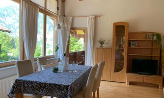 A bright dining room with a large table and chairs. In the background, there are windows overlooking nature and wooden paneling.