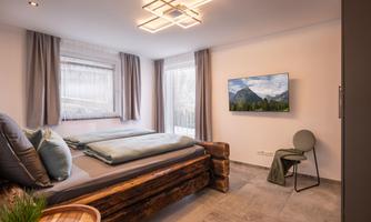 A modern bedroom with a large bed and a TV on the wall. The room has large windows that let in plenty of natural light.
