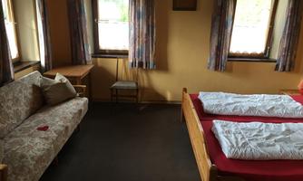 A simple room with two beds, a couch, and a table. The walls are painted in warm colors and there are several windows that let in daylight.