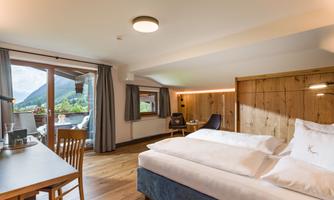 A modern hotel room with a large bed and wooden panels. There is a window with a view of nature and a desk.