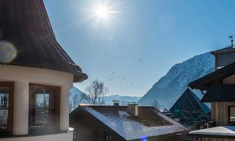 A sunny winter landscape with snow-covered roofs and mountains in the background. The sky is clear and blue.