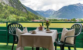 A beautiful balcony with a table on which a flower vase and plates are placed. In the background, majestic mountains and a green landscape can be seen.