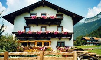 A traditional alpine house with many flower boxes and wooden cladding. In the background, mountains and a clear sky can be seen.