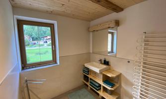 A modern bathroom with a wooden ceiling, a window, and a sink. There is a towel rack and stacked towels on a shelf.