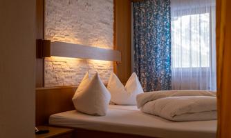 A cozy bedroom with a double bed, two pillows, and a bright, inviting atmosphere. The windows are covered with blue curtains and the room has a warm-lit wall.