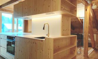 A modern kitchen made of light wood with a cooktop and sink. Large windows provide plenty of daylight.
