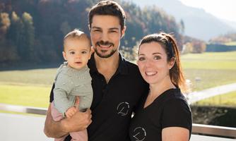 A family is smiling while posing outdoors. In the background, green meadows and mountains can be seen.
