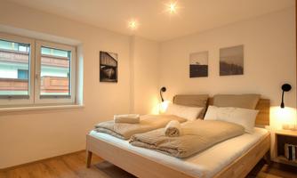 A cozy bedroom with a large double bed and soft lighting. Some pictures hang on the walls, and a window lets in daylight.