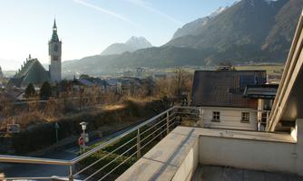 A picturesque view from a balcony overlooking the city of Schwaz with the parish church and mountains in the background. The sky is clear and the landscape is calm.