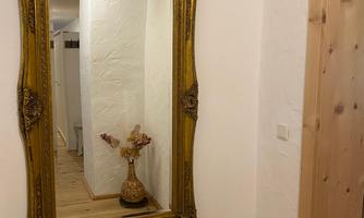 An elegant, gilded mirror hangs on the wall in a bright corridor. In the background, a decorative statue with flowers can be seen.