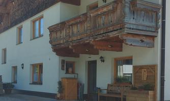 A traditional house with wooden balustrades and light-colored walls. In front of the house, there is a paved area with garden furniture.