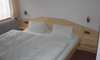 A simple bedroom with a double bed and two pillows. The walls are bright and there is a large window with curtains.