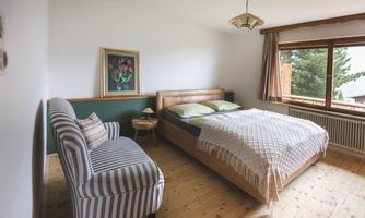 A cozy bedroom with a double bed and a comfortable seating area. Large windows offer a view of nature.