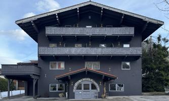A modern building in Alpine style with three balconies. The facade is dark gray and has a classic gable.