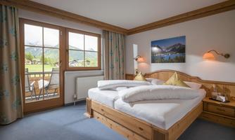 A cozy bedroom with a large double bed and wooden details. There is plenty of daylight coming through the windows and a view of the surroundings.