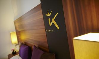 A stylish hotel room with a wooden bed back and a golden logo. Soft pillows and warm light create a cozy atmosphere.