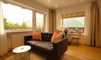 A modern living room with a gray sofa and colorful pillows. The view overlooks a terrace with a table and chairs, as well as a beautiful mountain landscape in the background.