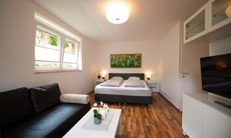A modern bedroom with two beds, a sofa, and a table. The walls are light, and there is a painting as well as a TV corner.