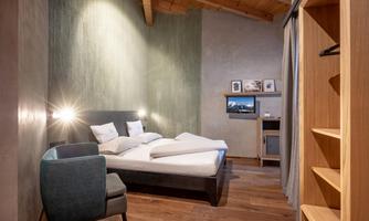 A modern hotel room with a double bed and an armchair. The walls are designed in soft colors and there is a television as well as a wooden shelf.