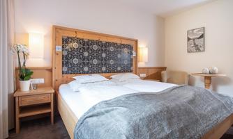 A comfortable bedroom with a large bed and a wooden frame. The walls are subtly decorated, and there is a cozy armchair as well as a small table.