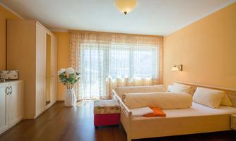 A bright bedroom with orange walls and wooden floor. There are two beds, a wardrobe, and large windows that let in plenty of daylight.