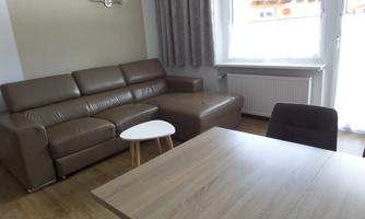 A modern furnished living room with a brown sofa and a small white table. There is a window with curtains and a wooden table in the room.