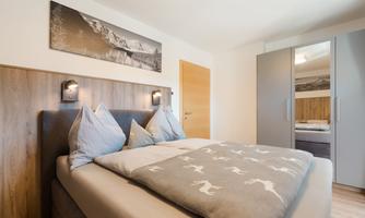A modern bedroom with a large bed and cozy pillows. A picture of mountains hangs on the wall, and a wardrobe is also visible.