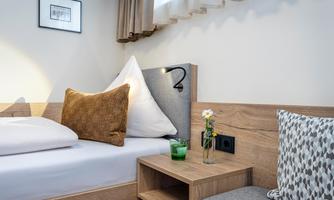 A cozy bedroom with a bed, pillows, and a wooden nightstand. On the nightstand, there is a glass with a flower and a decorative cushion.