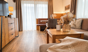 A modern living room with wooden details and cozy furnishings. The room is brightly lit by large windows with curtains.