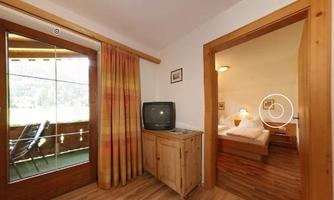 A cozy room with wooden flooring and a television. A balcony is visible through the window, and there is an adjoining sleeping area.