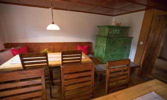 A cozy dining area with a large wooden table and wooden chairs. In the corner stands a green stove and there are red cushions on the bench.