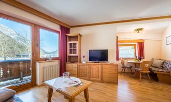 A cozy living room with wooden paneling and large windows. In the background, there is a dining table and a modern corner bench.
