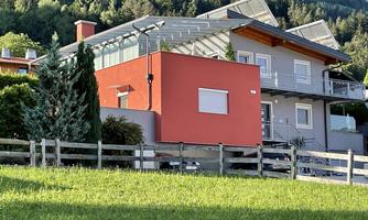 A modern house with a red facade and a spacious terrace area. The garden is well-kept and surrounded by a wooden fence.
