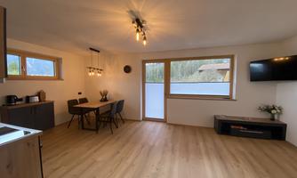 A modern living room with wooden floor and large windows. The room contains a dining table, a television, and a small kitchenette.