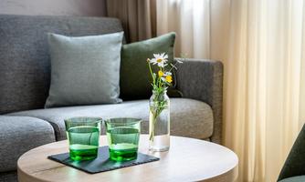 A cozy living area with a gray sofa and a round table. On the table are three green glasses and a small vase with flowers.