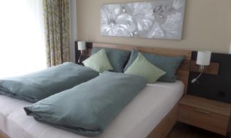 A modern bedroom with a double bed and green pillows. The wall is decorated with an elegant piece of artwork and a window with curtains.
