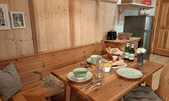 A cozy wooden dining table set with plates, glasses, and fresh flowers. In the background is the fully equipped kitchen area with a refrigerator and sideboard.