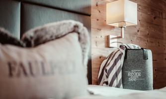 A cozy bedroom with a pillow and a wool blanket. Next to the bed, there is a small lamp on the wall.