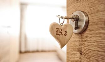 A heart-shaped keychain is hanging on a key in a door. The background shows a bright room with wooden details.