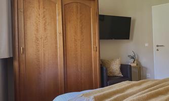 A cozy bedroom with a wooden wardrobe and a television on the wall. A seating area and decorative elements complete the space.