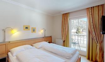 A cozy bedroom with two single beds and gentle lighting. Large windows overlooking a balcony create a bright and inviting atmosphere.