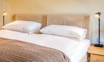 A cozy bedroom with a double bed and bright, soft pillows. Next to the bed are two modern bedside lamps.