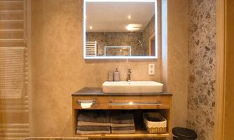 A modern bathroom with an elegant sink and flushable mirror lighting. Towels are neatly stacked on a shelf, and the floor is made of stone.