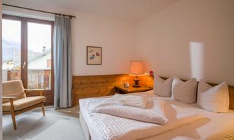 A cozy bedroom with a double bed, pillows, and a comfortable chair. Large windows let in plenty of daylight and provide a view outside.