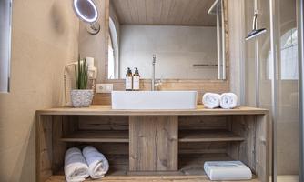 A modern bathroom with a wooden furniture piece and a white sink. Towels and toiletries are neatly arranged, and a large shower is visible.