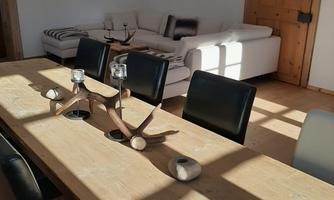 A bright, modern kitchen-living area with a large wooden table and black chairs. In the background, cozy sofas and some pictures on the wall can be seen.