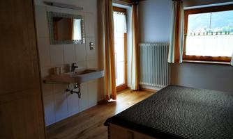 A simple bedroom with a bed, a sink, and a large window. The wooden details and the light curtains create a cozy atmosphere.