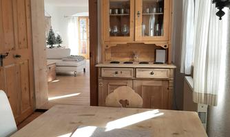A cozy kitchen with wooden furniture and a large dining table. Behind the table is a buffet with glass fronts, filled with some decorative items.